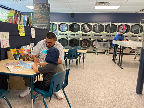 Wash and Learn library participants engage in table activity, and a row of coin laundry machines in the background.