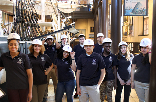 Group of students wearing hard hats, standing in front of museum exhibit.