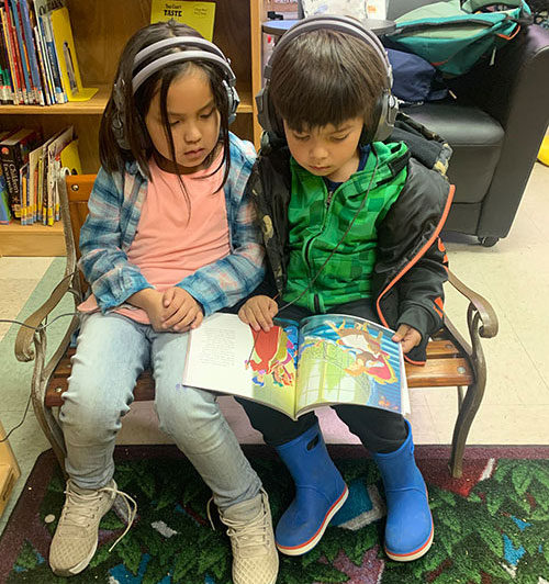 Two children sitting in chairs and reading a book.
