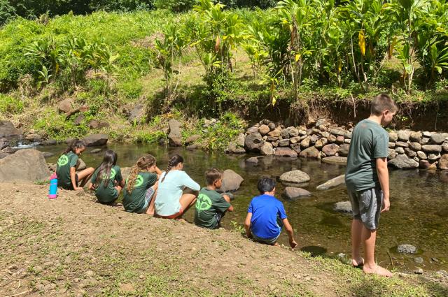 Children sit at the shore of a creek.