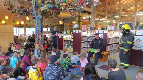 Two firefighters reading a book to a group of children in a library.